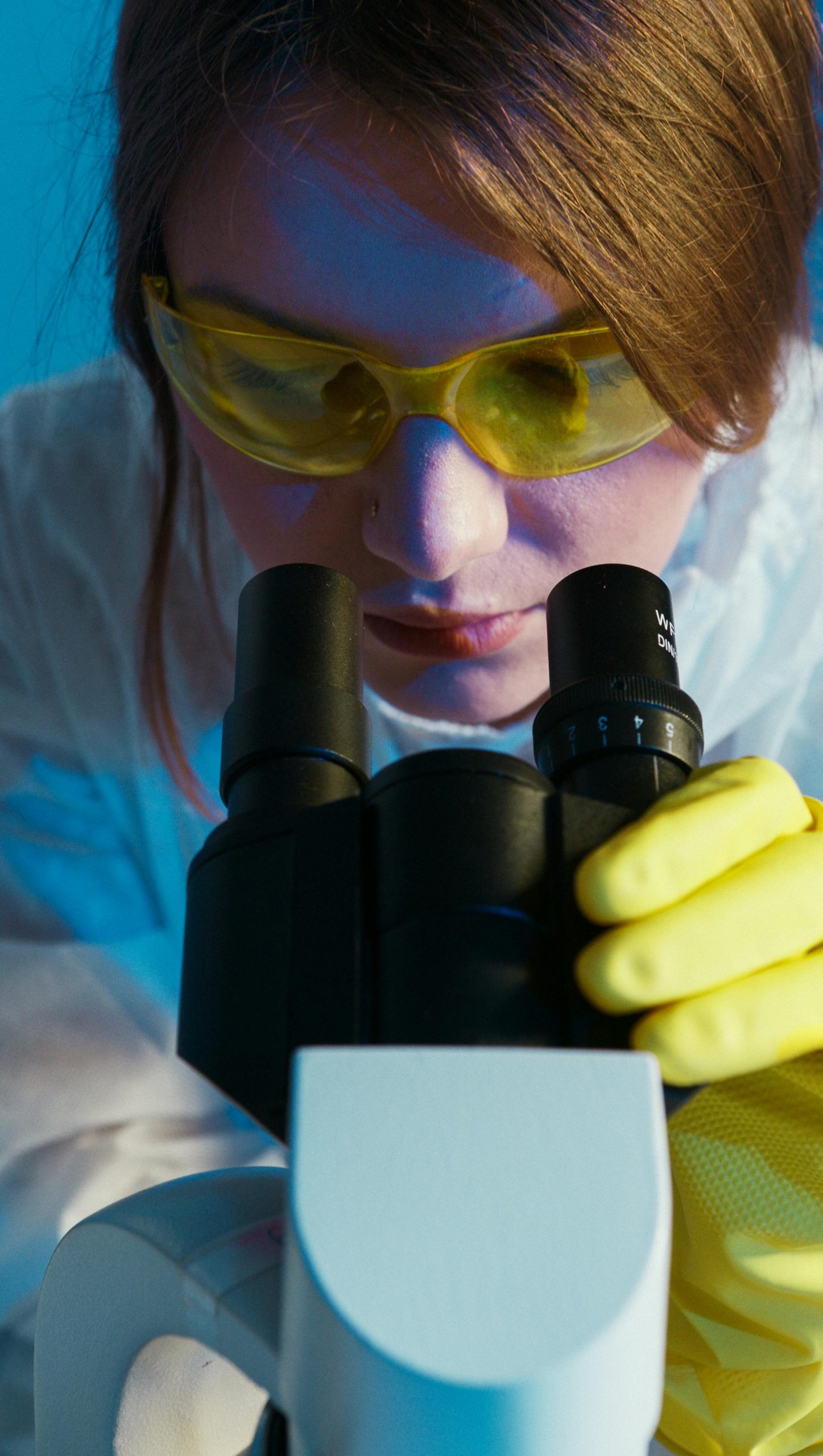 A female scientist in a lab coat looks through a microscope wearing yellow protective goggles and gloves.